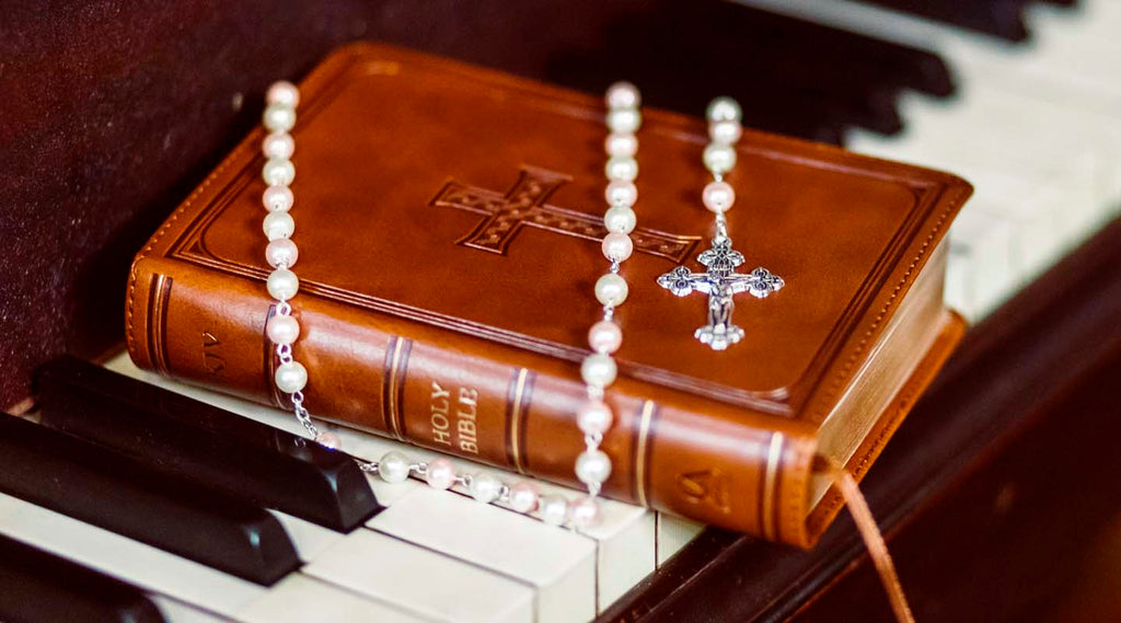 Pearl rosary with a silver crucifix draped over a leather-bound Holy Bible on piano keys, illustrating Catholic prayer and devotion.