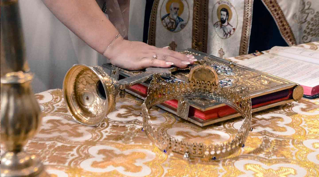 Hand placed on an iron cross and a Bible laying down on a table with a white and golden cloth.
