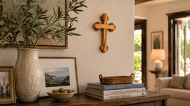 Olive wood cross hanging on a wall above a tabler with books, pictures and a vase with olive brunches