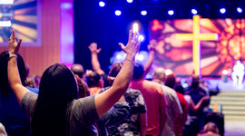 Christian worshippers raising their hands in prayer during a church service with a glowing cross at the front.
