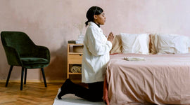 Woman kneeling and prating by the bed in a bedroom with a chair and nightstand.