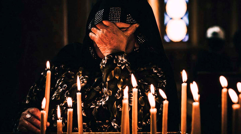Person in dark clothing praying with a hood sitting among lit candles in a dimly lit room.