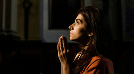 Woman in a brown outfit praying with hands pressed together in a dark setting