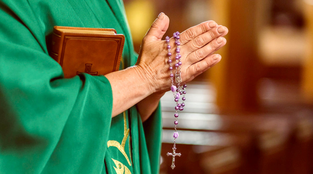 Priest in green robe holding a bible and prayinmg with purple rosary