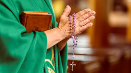 Priest in green robe holding a bible and prayinmg with purple rosary