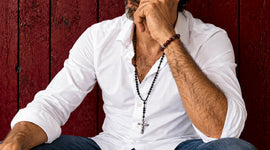 Man wearing a Catholic rosary necklace with black beads and a silver crucifix over a white shirt against a deep red background.