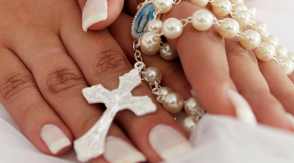 Hands holding a white pearl rosary with Miraculous Medal and silver crucifix during prayer or quiet reflection.