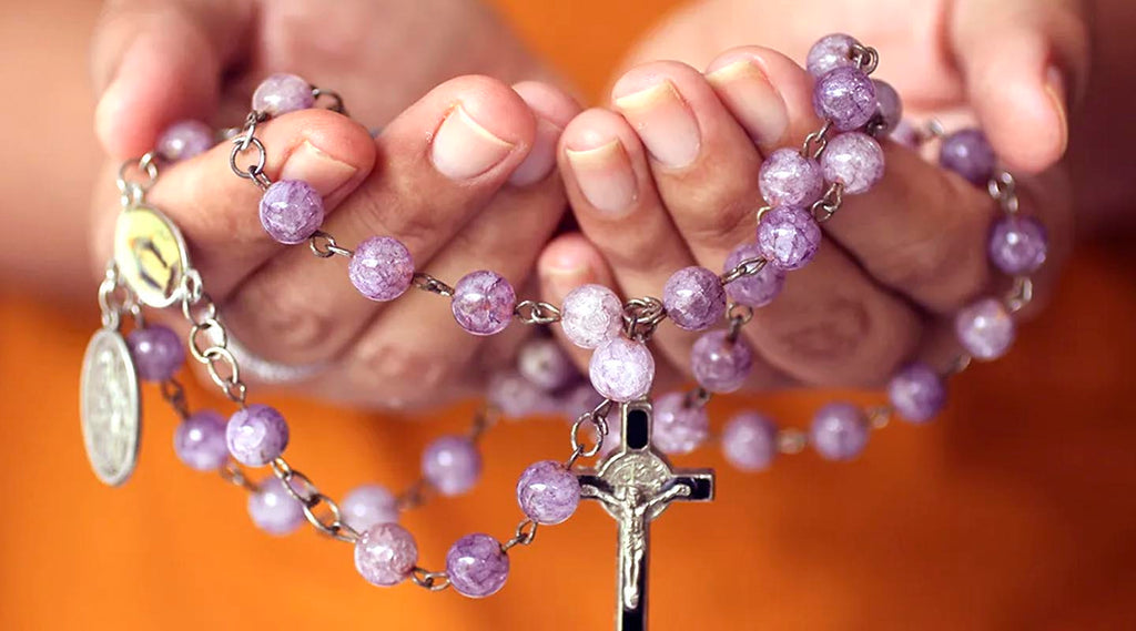 Purple rosary held in hands against a blurred orange background