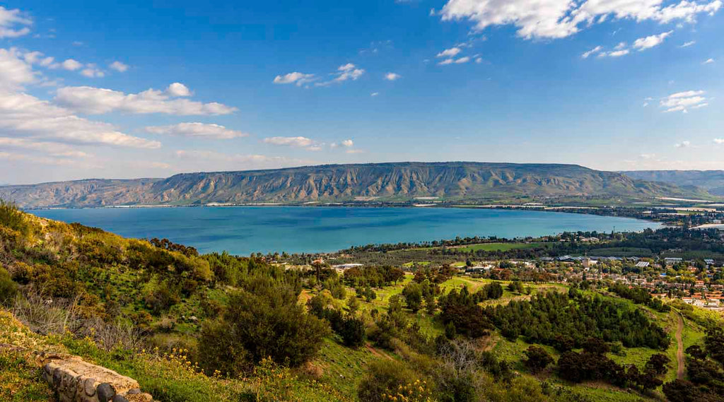 Panoramic view of the Sea of Galilee with lush hills, shoreline villages, and the Golan Heights under a blue sky.