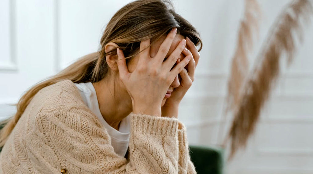 Woman in a beige sweater covering her face with both hands while experiencing emotional distress.
