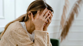 Woman in a beige sweater covering her face with both hands while experiencing emotional distress.