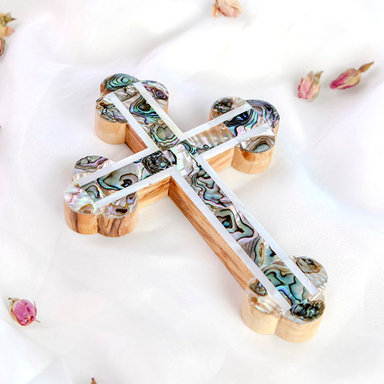 A decorative budded handmade olive wood cross with mother of pearl inlays, placed on a white surface with scattered flower petals around it.