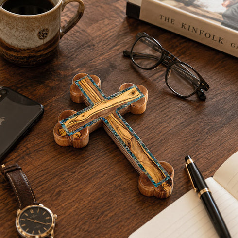 Wooden cross on a wooden desk with a cup, phone, glasses, and pen.