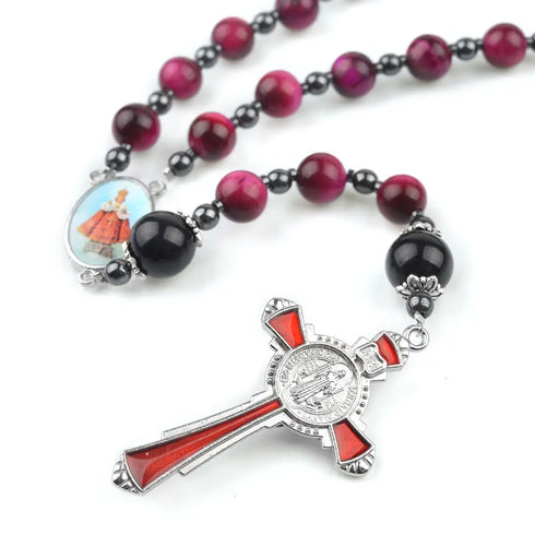 Handmade rosary with red Tiger Eye beads, a Child Jesus centrepiece and a red St. Benedict crucifix, laid on white surface.