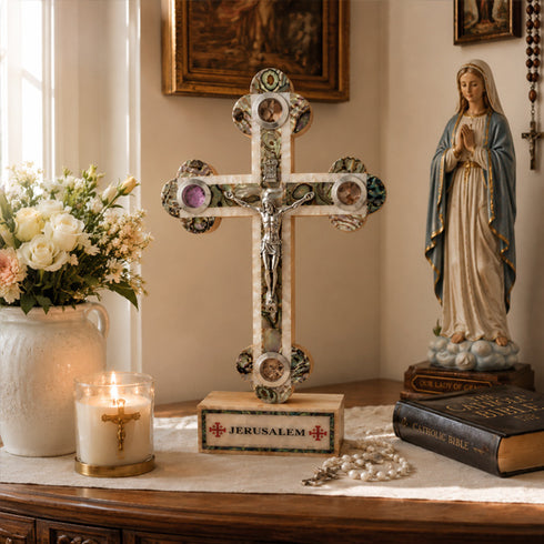 Decorative cross with Jerusalem inscription, candle, statue of a religious figure, and books on a table.