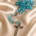 A rosary with translucent turquoise blue beads and a silver-tone metal crucifix, displayed on draped champagne satin fabric.