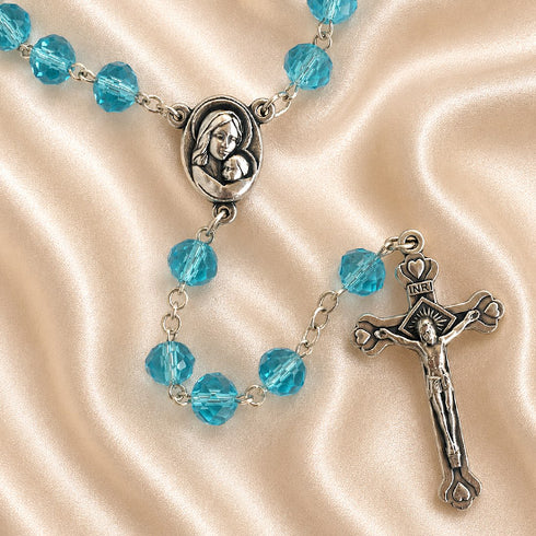 A rosary with light blue glass beads and silver-tone metal links, featuring a medallion of the Virgin Mary and a crucifix.