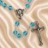 A rosary with light blue glass beads and silver-tone metal links, featuring a medallion of the Virgin Mary and a crucifix.