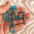 A close-up of a hand holding a rosary with turquoise-blue beads and a silver-tone crucifix, on a soft satin fabric background.