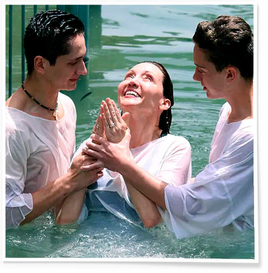 Woman dressed in white held by two priests, being happily baptised at the Jordan River.