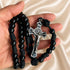 A black beaded rosary with a silver-toned crucifix held in a person's hand against a cream satin fabric background.