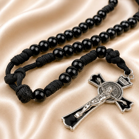 A black beaded rosary with a black cord and a silver crucifix, on a beige satin fabric background.