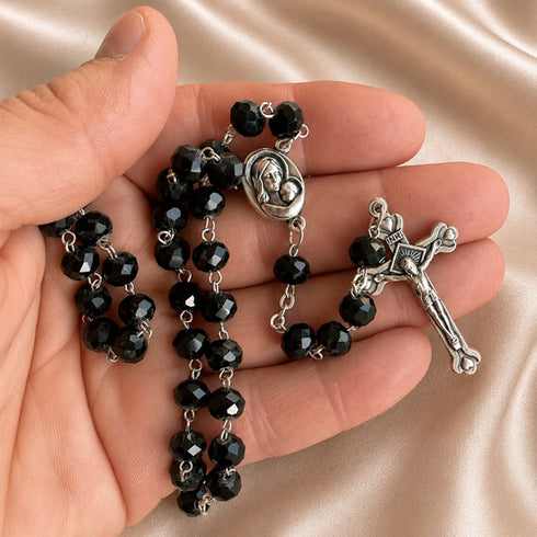 A close-up of a hand holding a black bead rosary with silver-tone metal links, a round medallion, and a crucifix pendant.