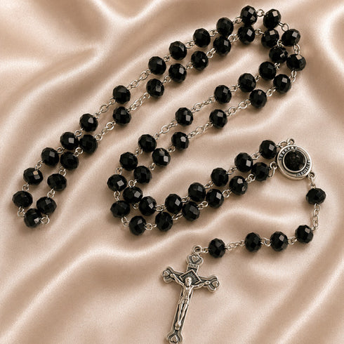 A black bead rosary with silver-tone chain links, a round centerpiece, and a silver crucifix, on a champagne fabric background.