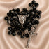 A black beaded rosary with silver-tone metal links, a medallion of the Virgin Mary, and a crucifix pendant, on a beige fabric background.