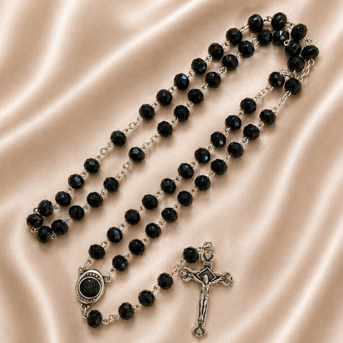 A black beaded rosary with silver-tone metal links, a medallion, and a crucifix, on a beige satin fabric background.