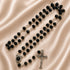 A black beaded rosary with silver-tone metal links, a medallion, and a crucifix, on a beige satin fabric background.