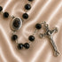 A close-up of a rosary with black beads, a silver-tone Jerusalem medallion and a silver crucifix pendant, on a satin background.