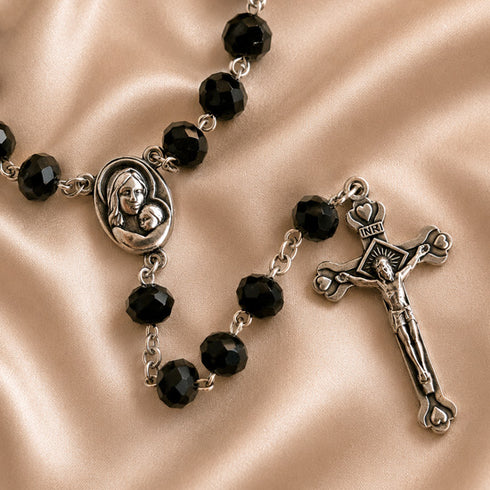 Black bead rosary with a silver-tone centerpiece medallion and crucifix, displayed on a satin beige background.