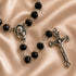 Black bead rosary with a silver-tone centerpiece medallion and crucifix, displayed on a satin beige background.