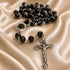 Black faceted bead rosary with a silver-tone crucifix and centerpiece, arranged on draped champagne satin fabric.