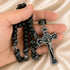 A close-up of a hand holding a black beaded rosary with black and white knotted cord and a silver-toned crucifix, on a satin fabric.