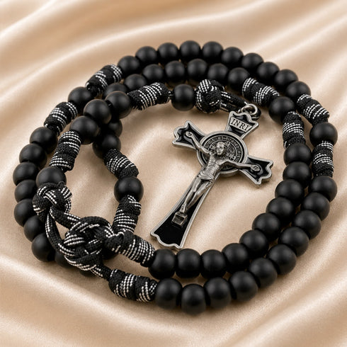 Black beaded rosary with a silver-toned crucifix and black-and-white knotted cord, on a satin fabric background.