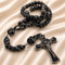 A black beaded rosary with black and white knotted cord accents and a black crucifix, on a beige satin fabric background.