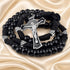 A black beaded rosary with a silver-tone crucifix, displayed on draped satin fabric.