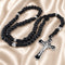 A black rosary with black cord and silver-tone metal accents, a crucifix, on a beige satin fabric background.