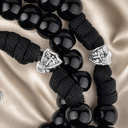 A close-up of a black rosary with glossy round beads, black cord, and two silver-toned shield-shaped charms featuring a cross design.