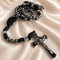 A gothic-style black and silver rosary with a crucifix, on a draped fabric background.