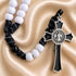 A rosary with white and black beads, blue cord, and a black-and-silver cross featuring a medallion on a beige background.
