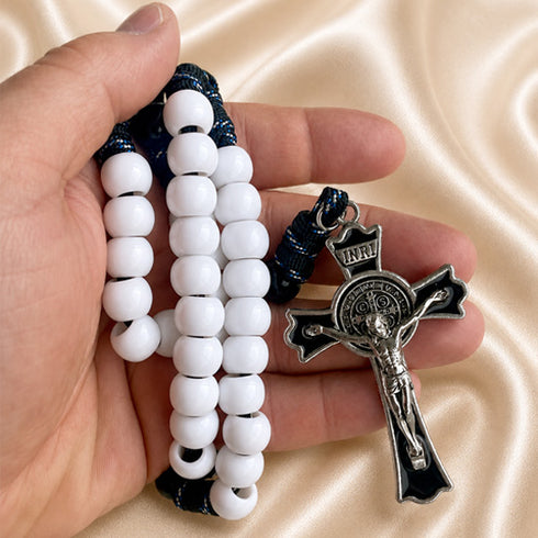 A hand holding a rosary with white beads, a dark blue cord, and a silver-toned crucifix on a beige fabric background.