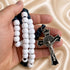 A hand holding a rosary with white beads, a dark blue cord, and a silver-toned crucifix on a beige fabric background.