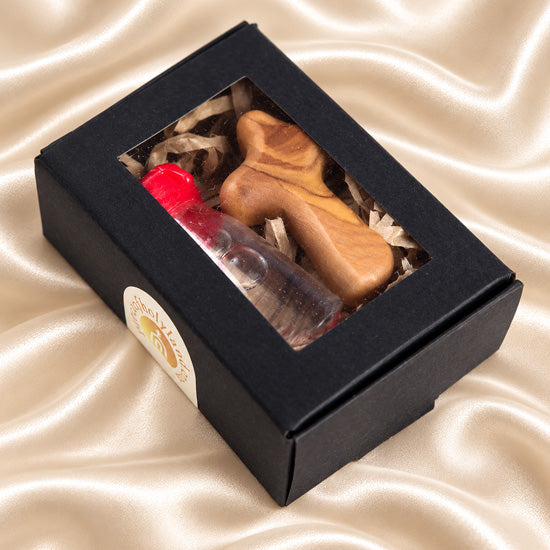 A black box containing a glass vial of Holy Water and an olive wood comfort cross, sealed with a red cap.
