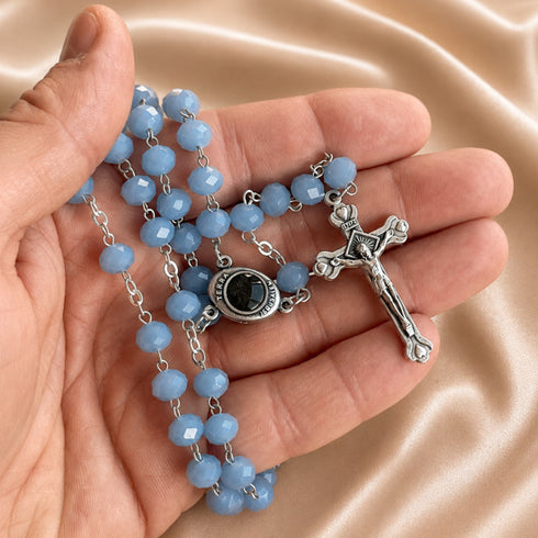 A close-up of a rosary necklace with light blue faceted beads, a silver crucifix, and a black centerpiece, held in a person's hand against satin background.