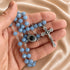 A close-up of a rosary necklace with light blue faceted beads, a silver crucifix, and a black centerpiece, held in a person's hand against satin background.