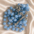 A light blue faceted bead rosary with silver-tone metal crucifix and medallion, on a beige satin fabric background.