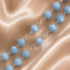 A close-up of a beaded chain necklace with light blue faceted round beads connected by silver-toned links, laid on a champagne satin fabric.